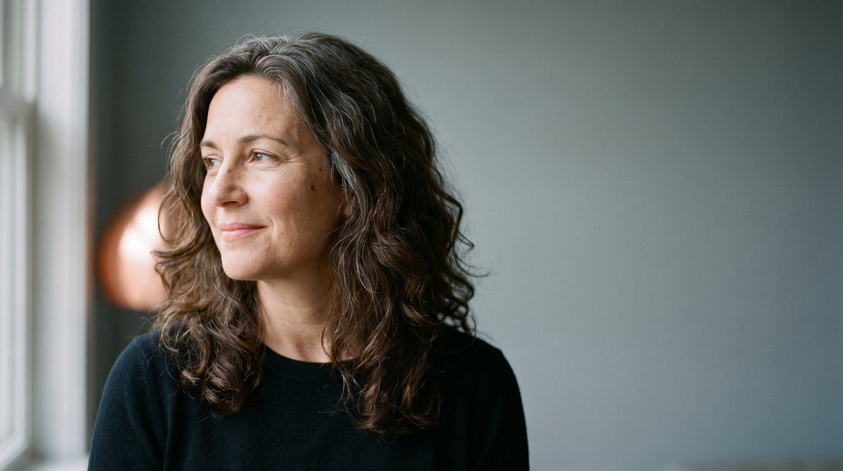A woman in her forties with thick, naturally curly dark hair, looking away from camera in soft window light